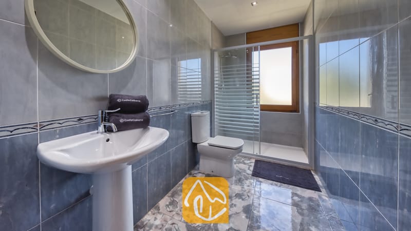 Ferienhäuser Costa Brava Spanien - Villa Baileys - Gefliestes Badezimmer mit Standwaschbecken, rundem Spiegel, WC und gläserner Duschkabine mit Milchglasfenster.