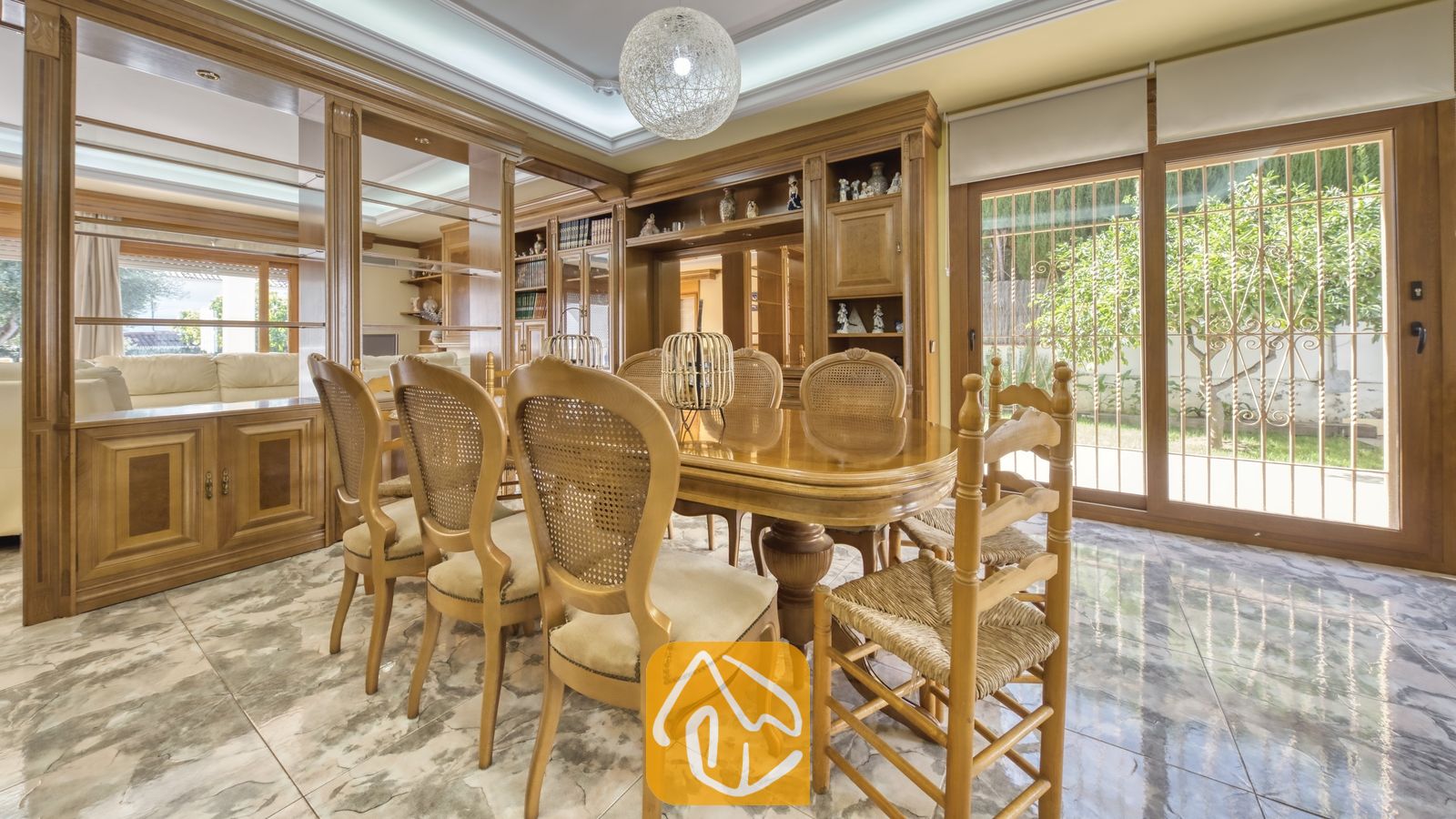 Vakantievilla's Costa Brava Spanje - Villa Baileys - Eetkamer met een gepolijste houten tafel, stoelen met rieten rugleuningen, inbouwkasten van hout, marmeren vloeren en grote glazen schuifpui die uitzicht biedt op een tuin.