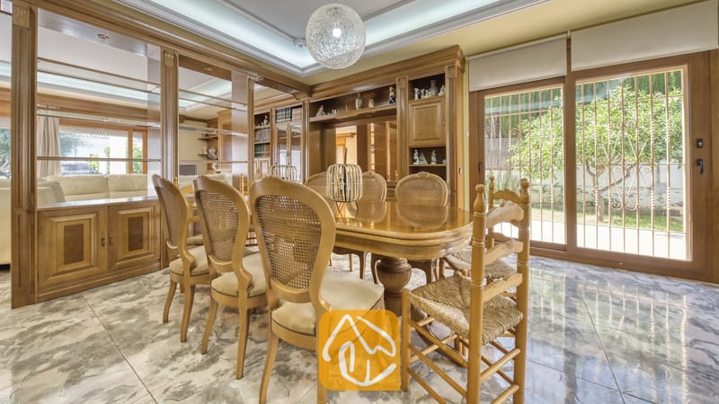 Vakantievilla's Costa Brava Spanje - Villa Baileys - Eetkamer met een gepolijste houten tafel, stoelen met rieten rugleuningen, inbouwkasten van hout, marmeren vloeren en grote glazen schuifpui die uitzicht biedt op een tuin.