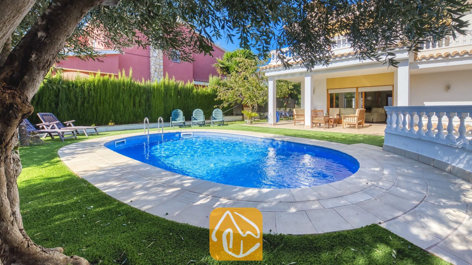 Vakantievilla's Costa Brava Spanje - Villa Baileys - Oval zwembad met blauw water en betegelde rand bij Villa Baileys, voorzien van ligstoelen op het gras en een overdekt terras op de achtergrond.