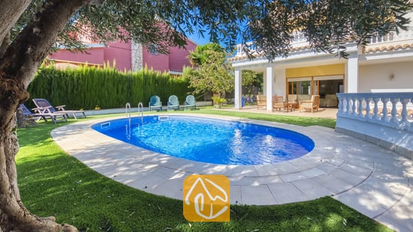 Vakantievilla's Costa Brava Spanje - Villa Baileys - Oval zwembad met blauw water en betegelde rand bij Villa Baileys, voorzien van ligstoelen op het gras en een overdekt terras op de achtergrond.