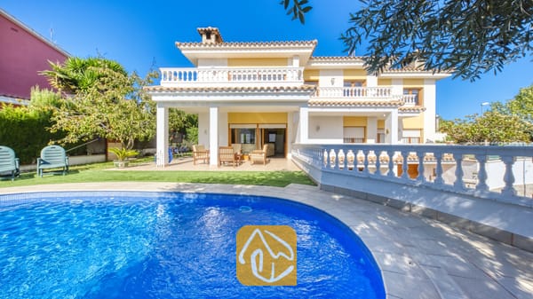 Vakantievilla's Costa Brava Spanje - Villa Baileys - Een gebogen, blauw getegeld zwembad op de voorgrond, met Villa Baileys en haar witte buitenkant, gele muren en betegelde balkons onder een heldere hemel.
