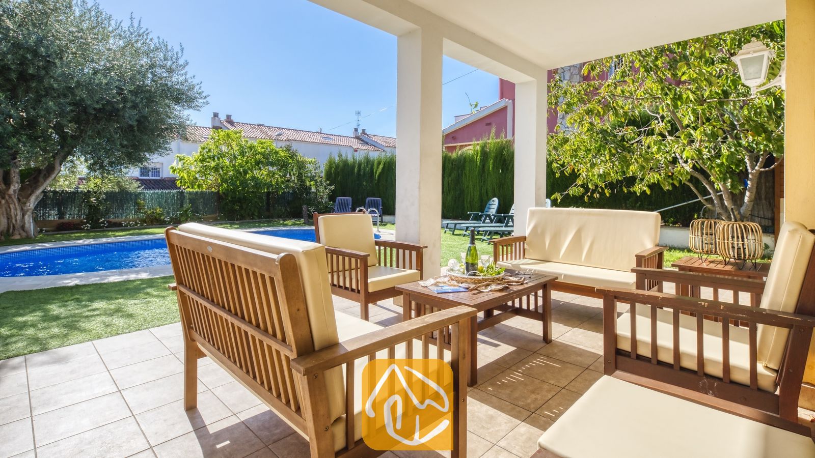 Vakantievilla's Costa Brava Spanje - Villa Baileys - Overdekt terras bij Villa Baileys met houten tuinmeubelen, crèmekleurige kussens en uitzicht op het zwembad en de tuin.