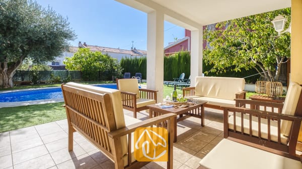 Vakantievilla's Costa Brava Spanje - Villa Baileys - Overdekt terras bij Villa Baileys met houten tuinmeubelen, crèmekleurige kussens en uitzicht op het zwembad en de tuin.