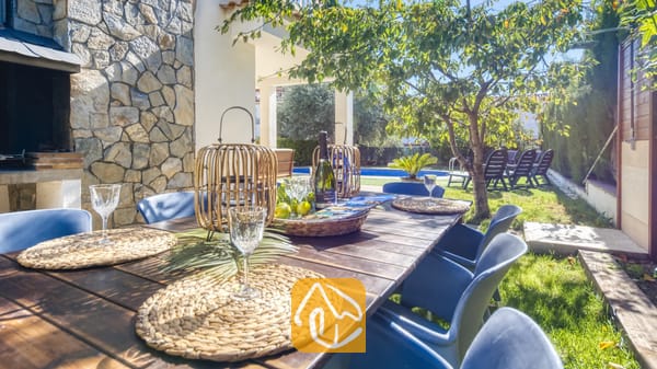 Vakantievilla's Costa Brava Spanje - Villa Baileys - Houten eettafel voor buiten, compleet met geweven placemats, wijnglazen en rotan lantaarns als middelpunt bij Villa Baileys in Spanje, met uitzicht op een tuin en zwembad.
