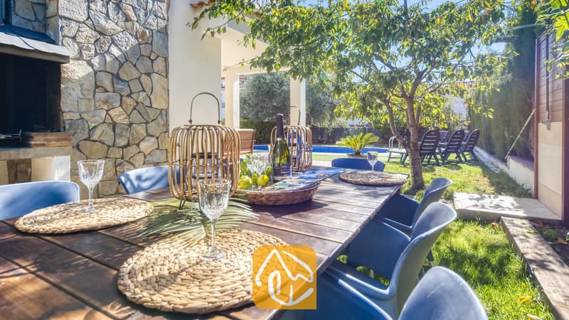 Vakantievilla's Costa Brava Spanje - Villa Baileys - Houten eettafel voor buiten, compleet met geweven placemats, wijnglazen en rotan lantaarns als middelpunt bij Villa Baileys in Spanje, met uitzicht op een tuin en zwembad.