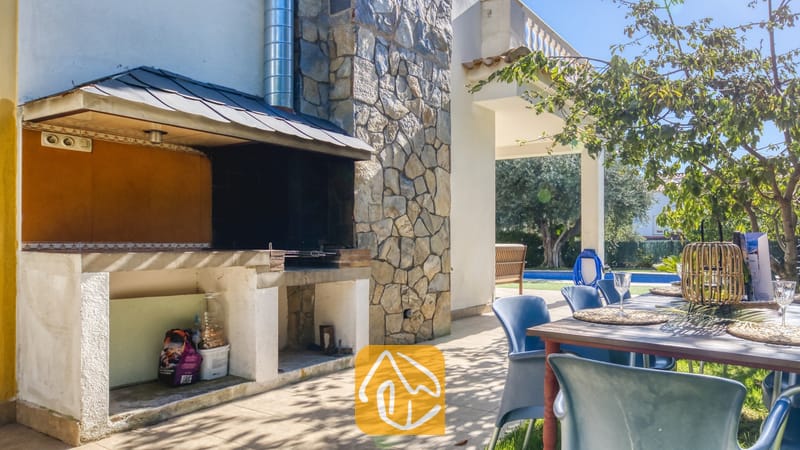 Vakantievilla's Costa Brava Spanje - Villa Baileys - Buitengedeelte met stenen barbecue bij Villa Baileys, voorzien van een stenen schoorsteen, een aangrenzend terras met eettafel en blauwe kunststof stoelen.