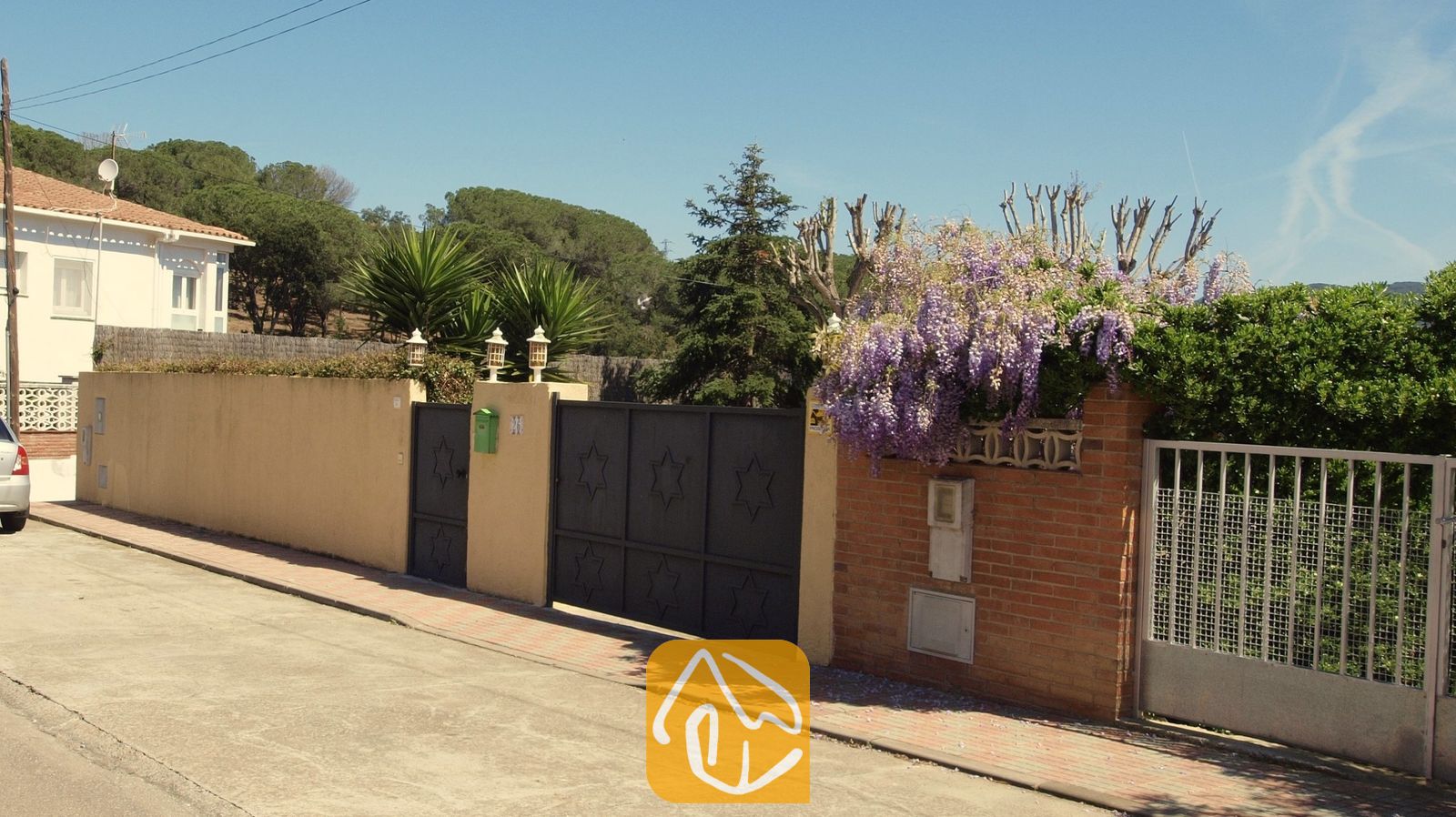 Vakantievilla's Costa Brava Spanje - Villa Miro - Zwarte metalen hekken met sterpatronen markeren de entree van Villa Miro, die wordt omringd door een gele muur en waar paarse blauweregen over de rode bakstenen hangt.
