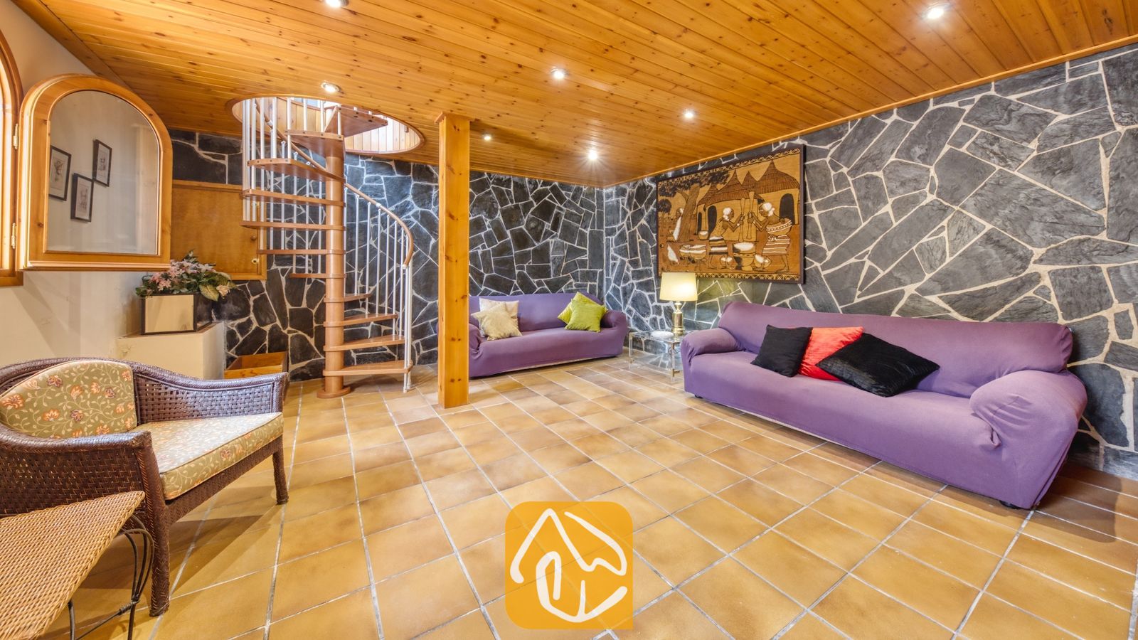 Vakantievilla's Costa Brava Spanje - Villa Miro - Woonkamer van Villa Miro met paarse banken, een houten wenteltrap en muren met een natuursteenlook. De terracotta plavuizen en het plafond met houten lambrisering maken het rustieke interieur compleet.