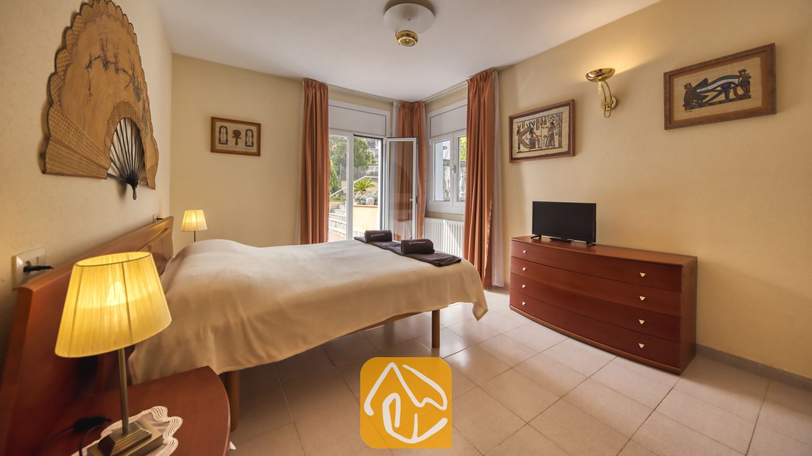 Vakantievilla's Costa Brava Spanje - Villa Miro - Slaapkamer in Villa Miro, voorzien van een tweepersoonsbed met een licht sprei, een houten dressoir met televisie en een grote decoratieve waaier aan de muur boven het hoofdeinde.