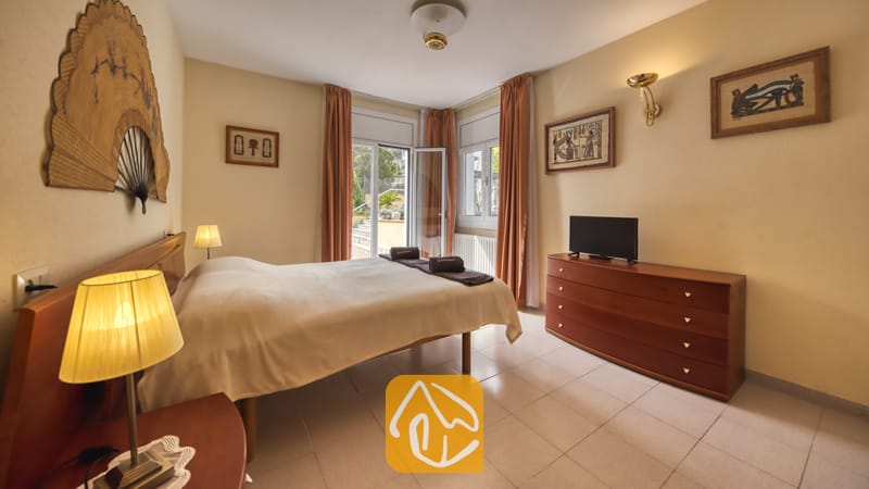 Villas de vacances Costa Brava Espagne - Villa Miro - Chambre de la Villa Miro équipée d'un lit double avec un couvre-lit léger, d'une commode en bois avec télévision, et d'un grand éventail décoratif fixé au mur au-dessus de la tête de lit.