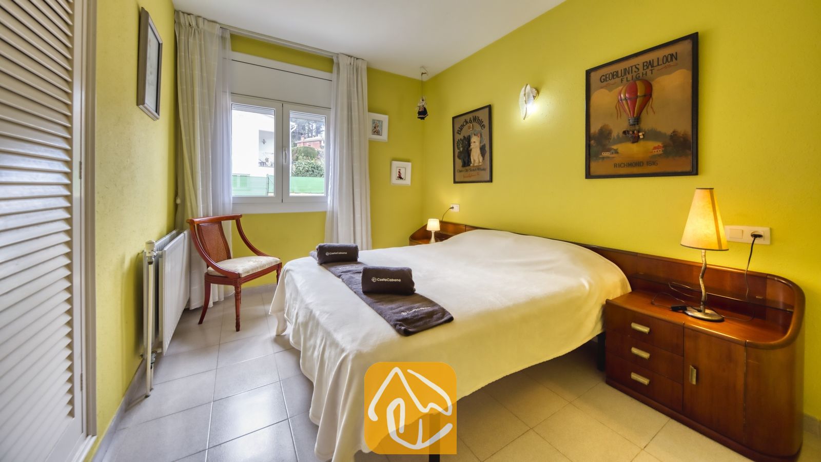 Vakantievilla's Costa Brava Spanje - Villa Miro - Slaapkamer met gele muren in Villa Miro, voorzien van een tweepersoonsbed met wit beddengoed, houten nachtkastjes en ingelijste kunstwerken boven het hoofdeinde.