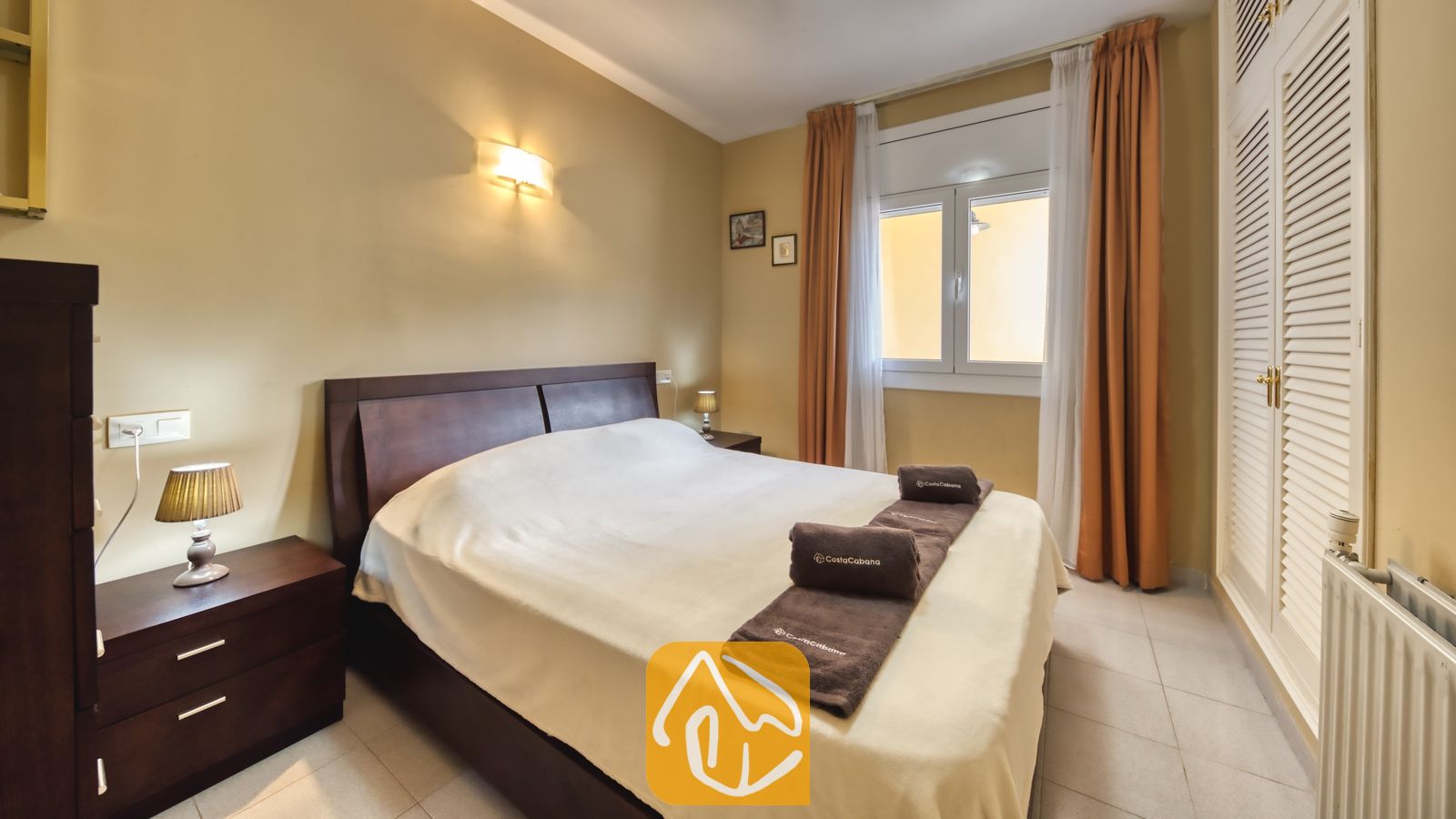Vakantievilla's Costa Brava Spanje - Villa Miro - Slaapkamer in Villa Miro, voorzien van een tweepersoonsbed van donker hout, bijpassende nachtkastjes met lampen en oranje gordijnen die het raam omlijsten.
