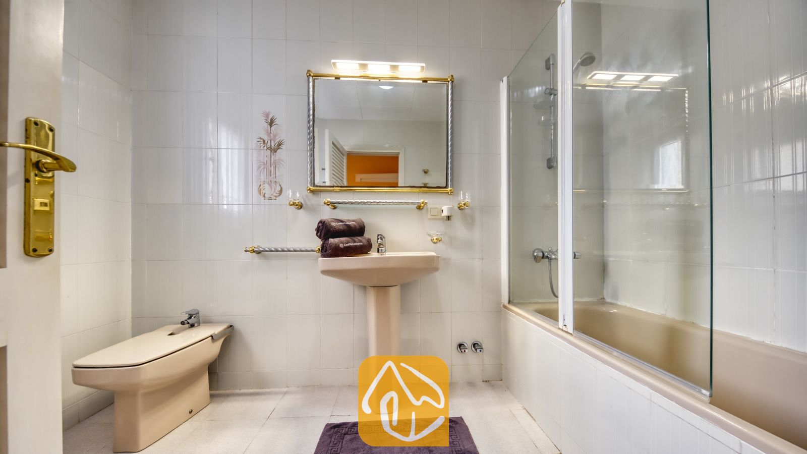 Vakantievilla's Costa Brava Spanje - Villa Miro - Badkamer voorzien van een beige wastafel op zuil, bidet en een ligbad met glazen douchescherm. De wit betegelde wanden worden geaccentueerd door goudkleurige armaturen en een verlichte spiegel boven de wastafel.