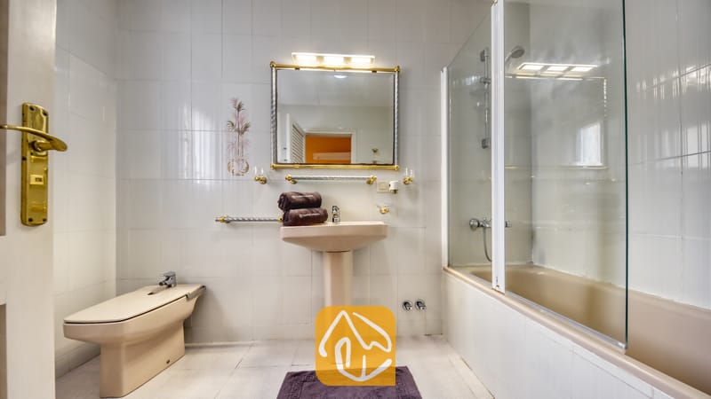 Villas de vacances Costa Brava Espagne - Villa Miro - Salle de bain équipée d'un lavabo sur colonne beige, d'un bidet et d'une baignoire avec pare-douche vitré. Les murs en carrelage blanc sont rehaussés par des finitions dorées et un miroir éclairé surplombant le lavabo.