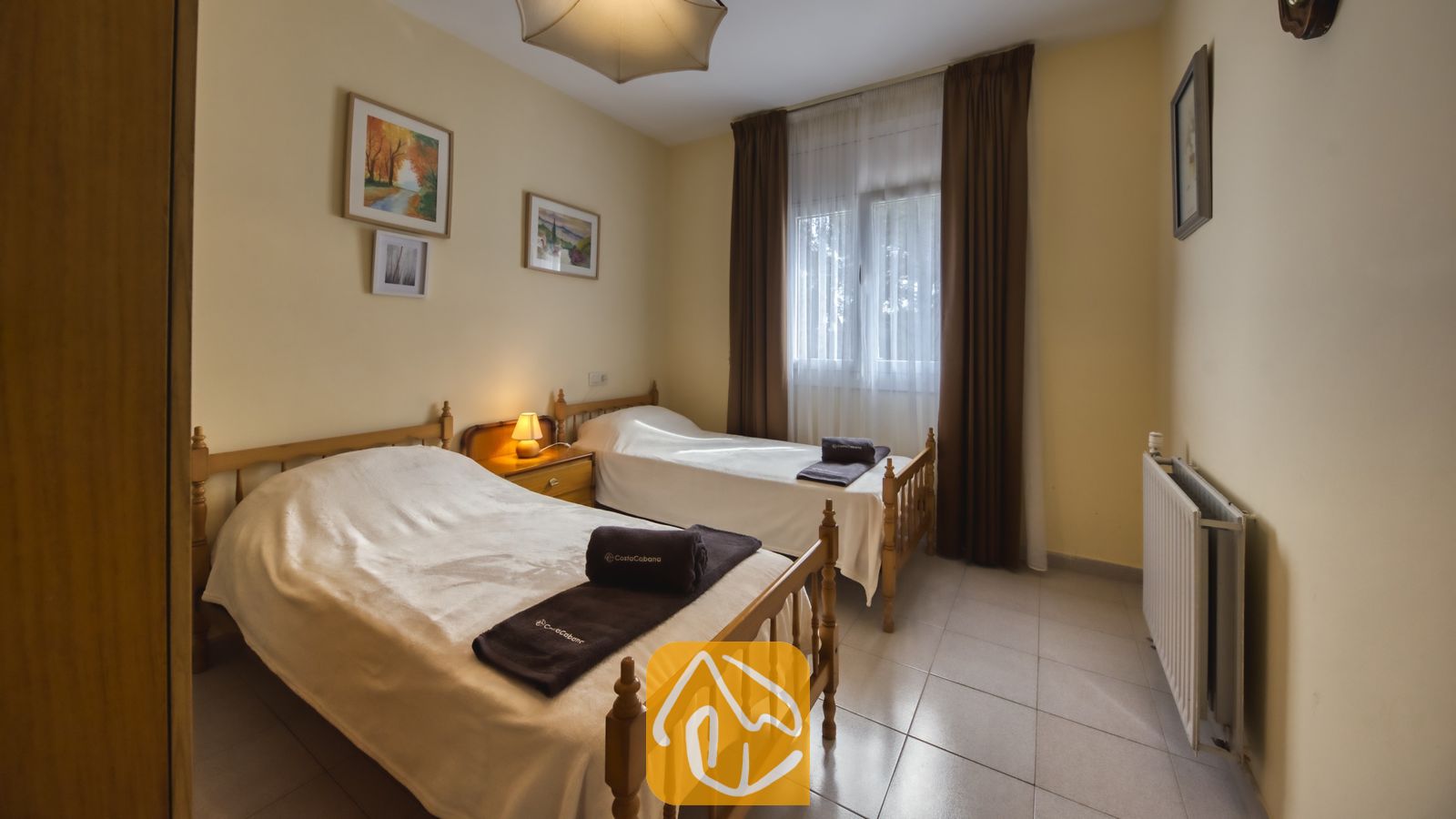 Vakantievilla's Costa Brava Spanje - Villa Miro - Slaapkamer met twee houten eenpersoonsbedden in Villa Miro, voorzien van wit beddengoed, donkerbruine handdoeken, een bedlampje en een witte radiator.