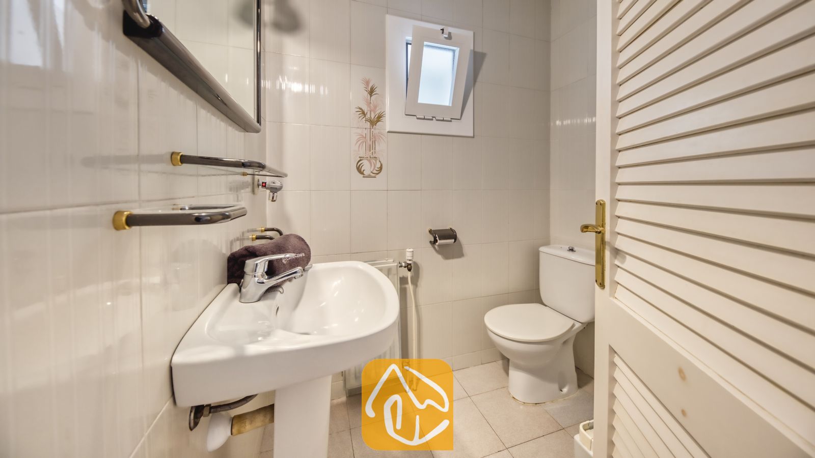 Vakantievilla's Costa Brava Spanje - Villa Miro - Badkamer voorzien van een witte wastafel op zuil, toilet en betegelde wanden. Boven het toilet bevindt zich een klein kiepraam met matglas, naast een witte louvredeur.