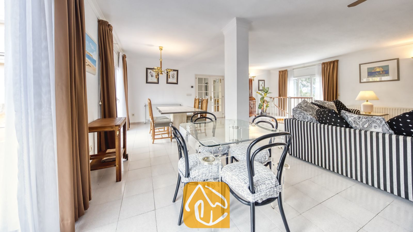 Vakantievilla's Costa Brava Spanje - Villa Miro - Glazen eettafel met zwarte stoelen in een open woonruimte bij Villa Miro, voorzien van een gestreepte bank en witte plavuizenvloeren.