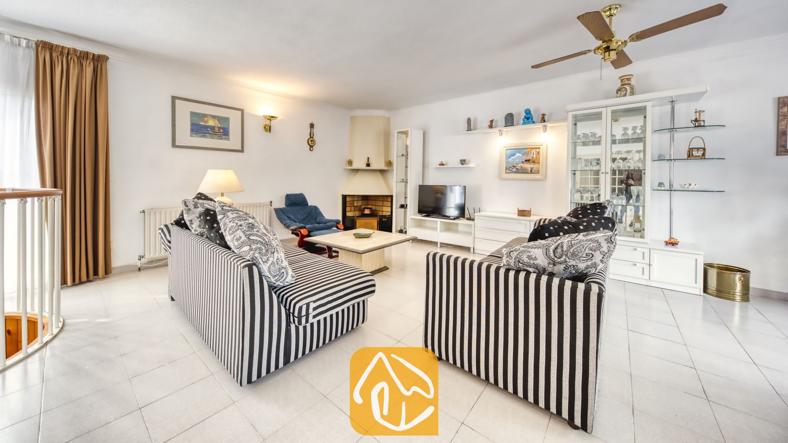 Vakantievilla's Costa Brava Spanje - Villa Miro - De woonkamer van Villa Miro is voorzien van zwart-wit gestreepte banken, een hoekhaard en een houten plafondventilator. De witte plavuizenvloer en een glazen vitrinekast maken het geheel compleet.