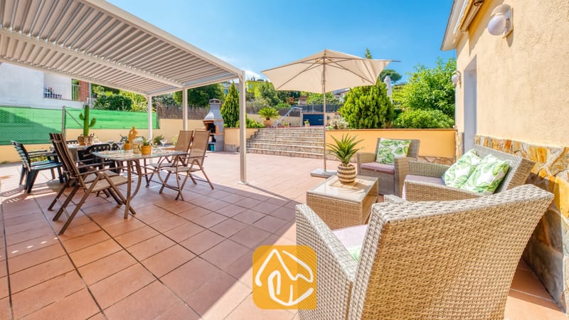 Vakantievilla's Costa Brava Spanje - Villa Miro - Op het betegelde terras van Villa Miro staat een wicker loungeset met kussens in een bladerdessin, vergezeld door een eettafel onder een pergola en een stenen barbecue.