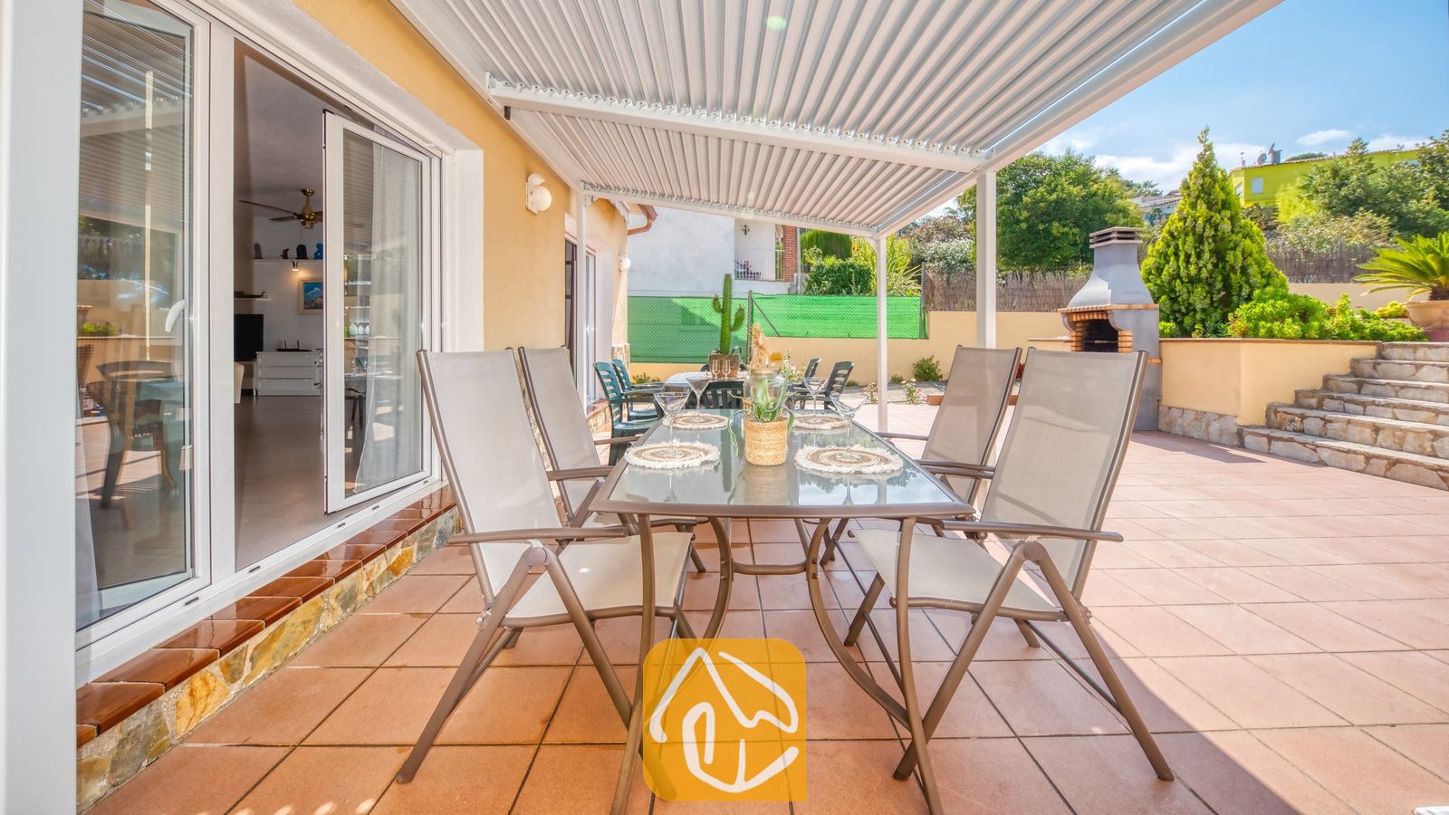 Vakantievilla's Costa Brava Spanje - Villa Miro - Overdekt terras bij Villa Miro met een glazen eettafel, moderne grijze stoelen en een ingebouwde stenen barbecue op een betegeld terras.