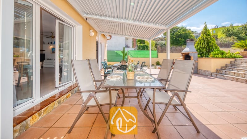 Vakantievilla's Costa Brava Spanje - Villa Miro - Overdekt terras bij Villa Miro met een glazen eettafel, moderne grijze stoelen en een ingebouwde stenen barbecue op een betegeld terras.
