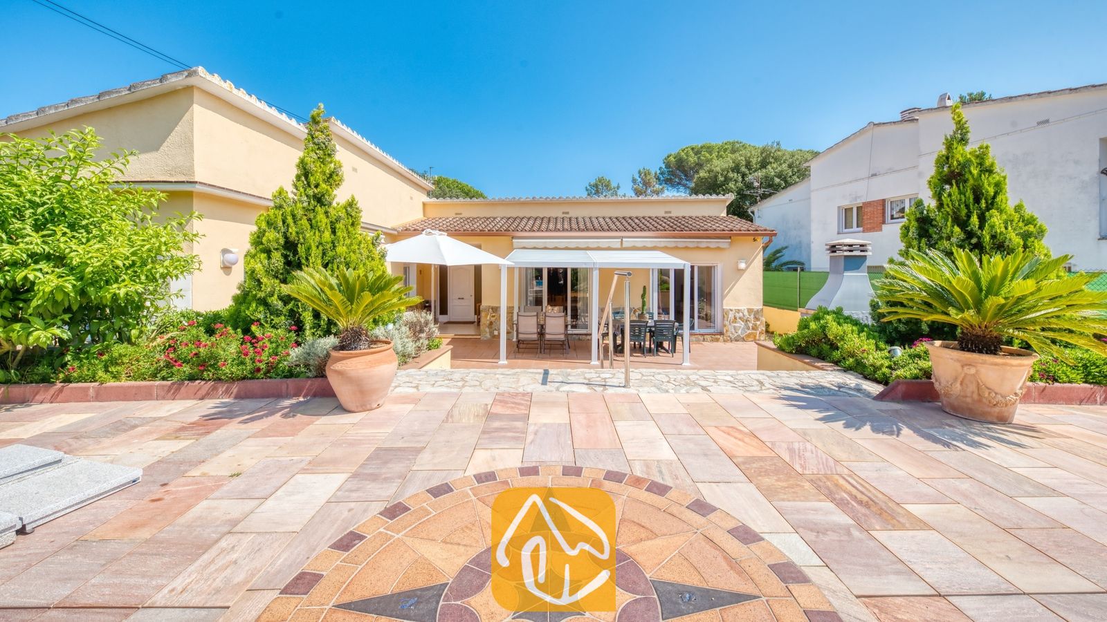 Vakantievilla's Costa Brava Spanje - Villa Miro - Terras van Villa Miro met een stenen kompasmozaïek, buitenmeubilair onder een witte pergola en palmbomen in potten tegen de gele gevel van het huis.