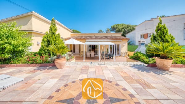 Vakantievilla's Costa Brava Spanje - Villa Miro - Terras van Villa Miro met een stenen kompasmozaïek, buitenmeubilair onder een witte pergola en palmbomen in potten tegen de gele gevel van het huis.