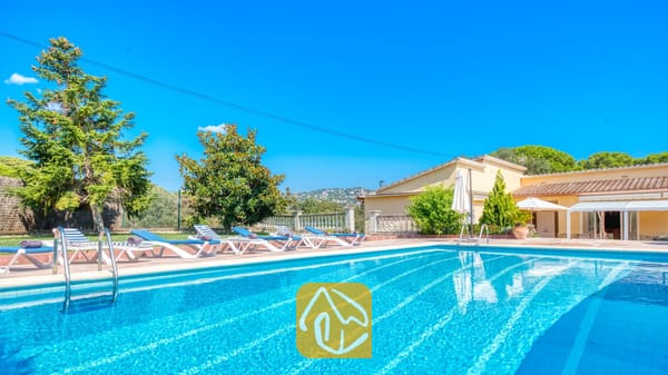Vakantievilla's Costa Brava Spanje - Villa Miro - Buitenzwembad bij Villa Miro met blauw-witte ligbedden op een terras van terracotta. Groene bomen en een geel gebouw onder een strakblauwe lucht.