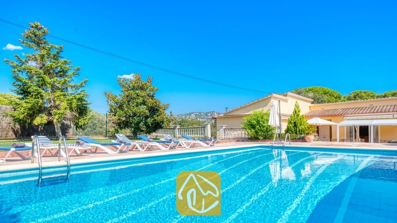 Vakantievilla's Costa Brava Spanje - Villa Miro - Buitenzwembad bij Villa Miro met blauw-witte ligbedden op een terras van terracotta. Groene bomen en een geel gebouw onder een strakblauwe lucht.
