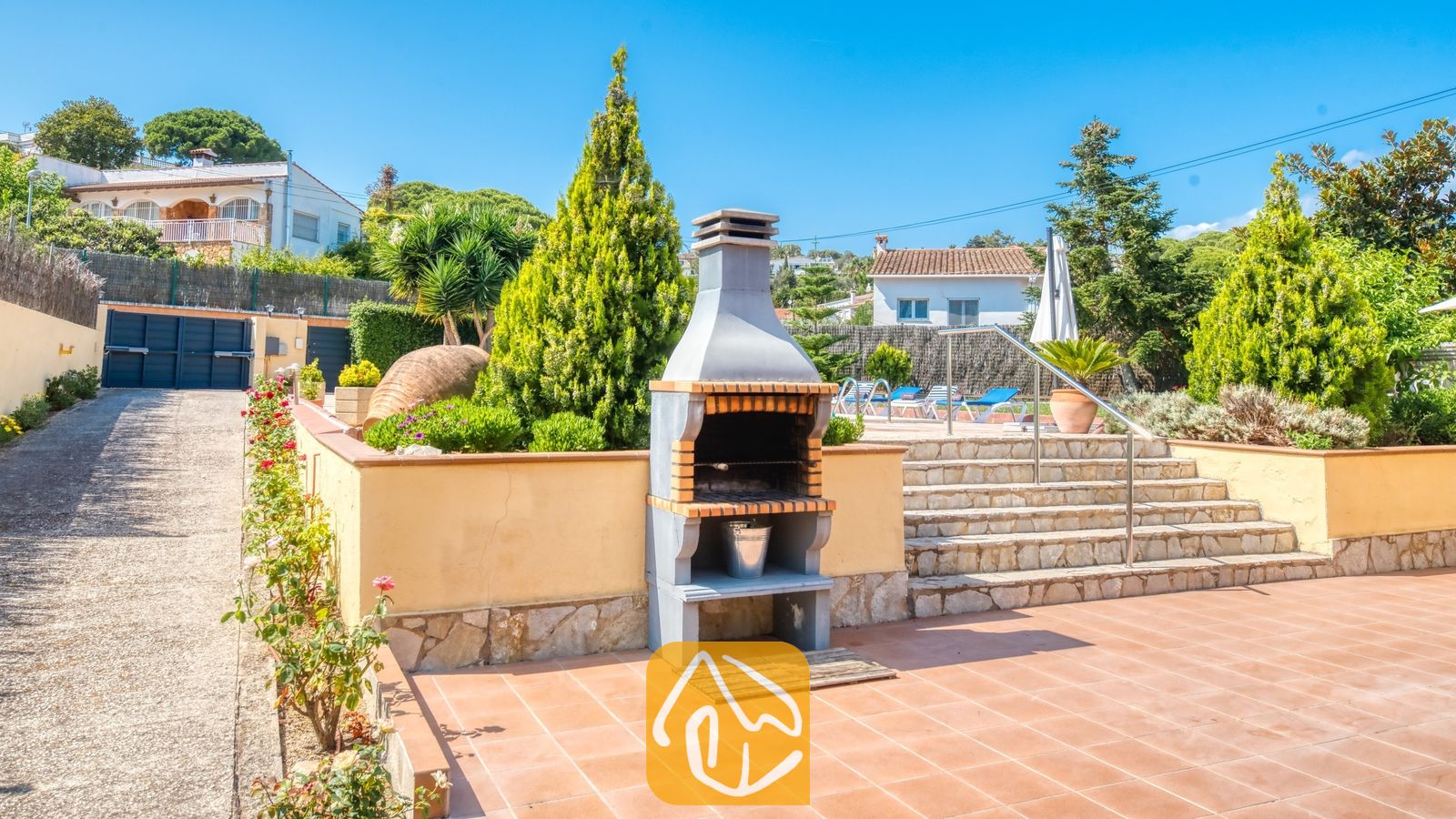 Vakantievilla's Costa Brava Spanje - Villa Miro - Stenen barbecue op een terras van terracotta met een trap die naar het zwembad leidt. Villa Miro beschikt over aangelegde tuinen met coniferen en een metalen leuning.