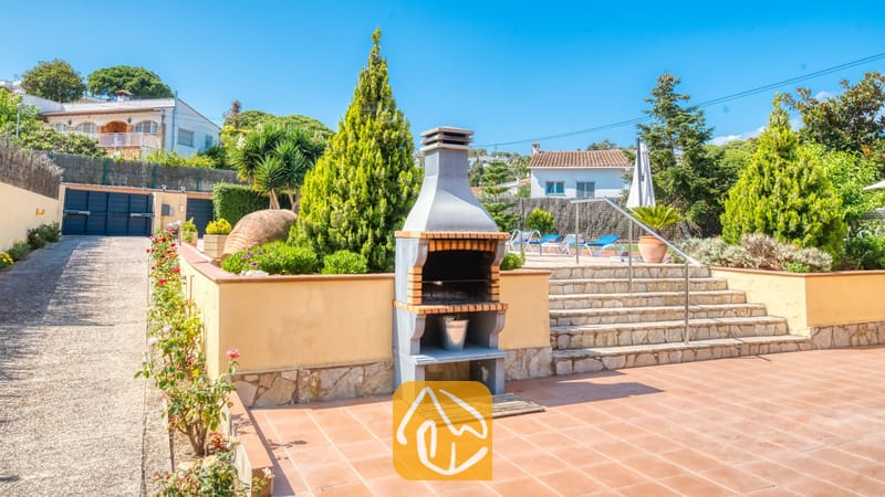 Vakantievilla's Costa Brava Spanje - Villa Miro - Stenen barbecue op een terras van terracotta met een trap die naar het zwembad leidt. Villa Miro beschikt over aangelegde tuinen met coniferen en een metalen leuning.
