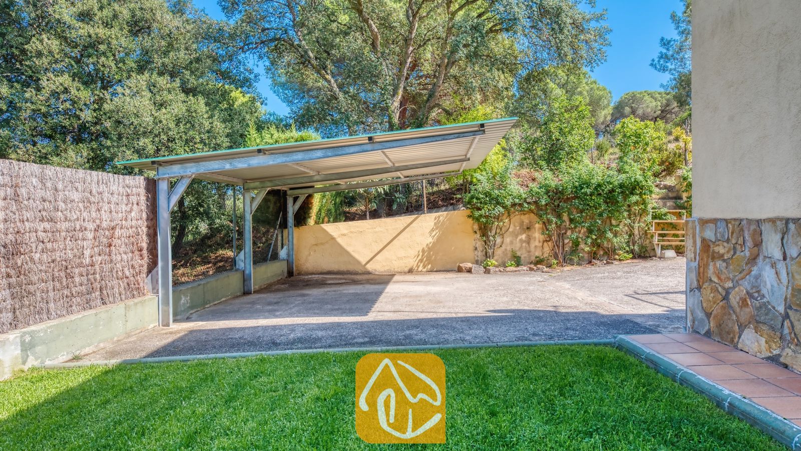 Vakantievilla's Costa Brava Spanje - Villa Miro - De metalen carport bij Villa Miro biedt een schaduwrijke parkeerplaats op een betonnen ondergrond, omzoomd door een rieten schutting, een groen gazon en weelderige mediterrane bomen.