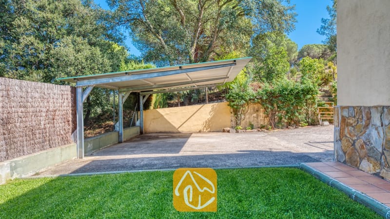 Villas de vacances Costa Brava Espagne - Villa Miro - L'abri voiture métallique de la Villa Miro offre un stationnement ombragé sur une dalle en béton, bordé par une clôture en roseaux, une pelouse verdoyante et des arbres méditerranéens luxuriants.