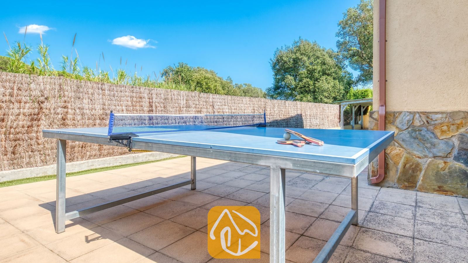 Vakantievilla's Costa Brava Spanje - Villa Miro - Tafeltennistafel met een blauw blad en batjes op een betegeld terras. Een rieten schutting en een stenen muur vormen de afscheiding van de speelruimte buiten bij Villa Miro.