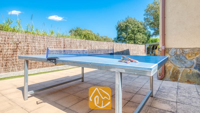 Villas de vacances Costa Brava Espagne - Villa Miro - Une table de ping-pong au plateau bleu, accompagnée de ses raquettes, est installée sur une terrasse carrelée. Une clôture en roseaux et un mur en pierre délimitent l'espace de jeu extérieur de la Villa Miro.