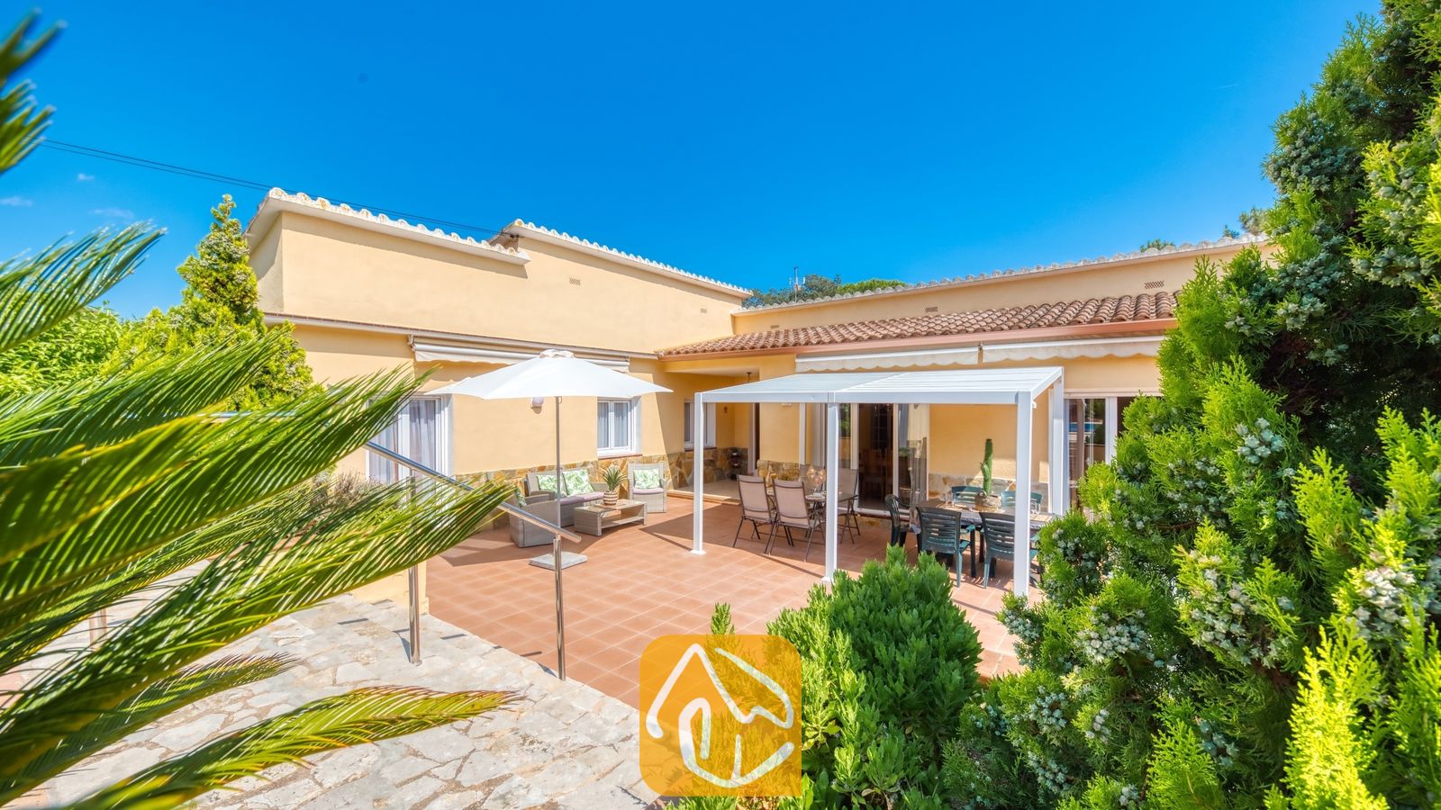 Vakantievilla's Costa Brava Spanje - Villa Miro - Terracottaterras bij Villa Miro met een witte pergola, buitenmeubilair om te dineren en een aparte zithoek met parasol, omringd door groen struikgewas.