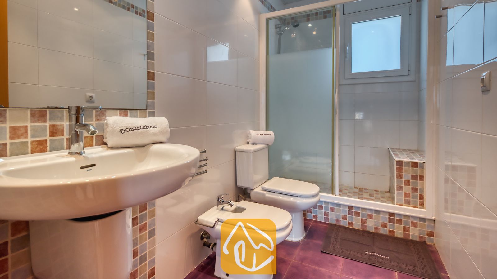 Vakantievilla's Costa Brava Spanje - Villa Sandy Beach - Badkamer met een witte wastafel op zuil, bidet en toilet. De inloopdouche is voorzien van aardetinten mozaïektegels en een glazen deur, gecombineerd met paarse vloertegels.