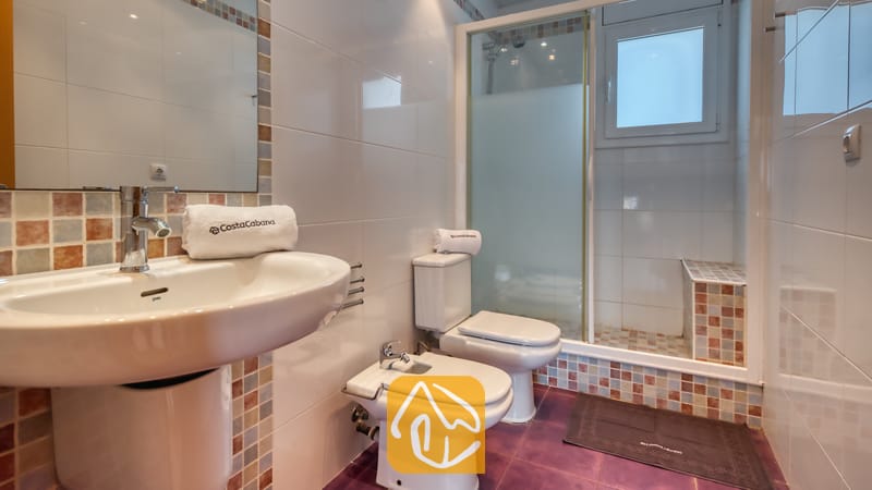 Villas de vacances Costa Brava Espagne - Villa Sandy Beach - Salle de bain équipée d'un lavabo sur colonne blanc, d'un bidet et de toilettes. La douche à l'italienne est agrémentée de mosaïques aux tons naturels et d'une porte vitrée, le tout contrastant avec un carrelage au sol violet.