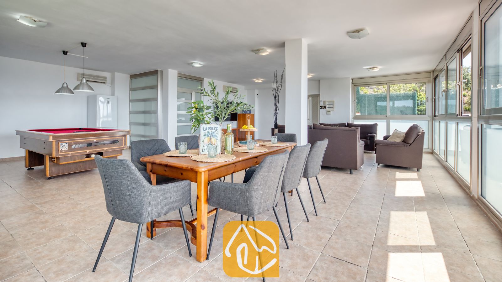 Vakantievilla's Costa Brava Spanje - Villa Sandy Beach - Houten eettafel met grijze stoffen stoelen in een open ruimte bij Villa Sandy Beach, voorzien van een biljarttafel met rood laken en bruine banken bij grote raampartijen.