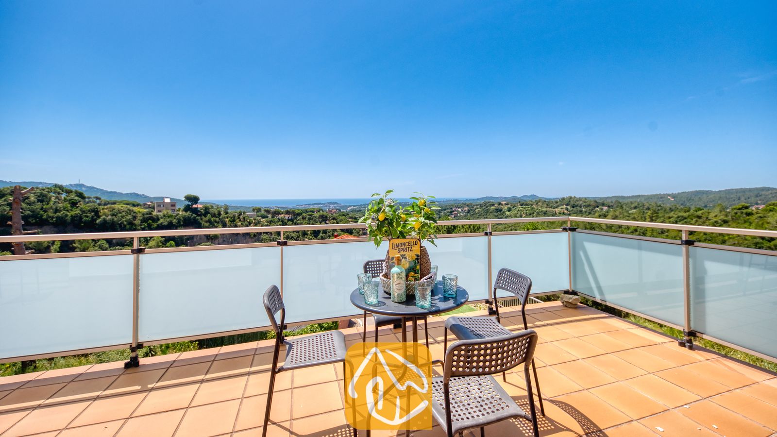 Vakantievilla's Costa Brava Spanje - Villa Sandy Beach - Terras met uitzicht op de groene heuvels en de zee bij Villa Sandy Beach. Een zwarte tafel met drankjes en een citroenboom staan op de terracotta tegels, omringd door een glazen balustrade.