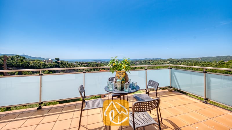 Villas de vacances Costa Brava Espagne - Villa Sandy Beach - Terrasse avec vue sur les collines verdoyantes et la mer à la Villa Sandy Beach. Une table noire accompagnée de boissons et un citronnier agrémentent le sol en terre cuite, le tout entouré d'une balustrade en verre.