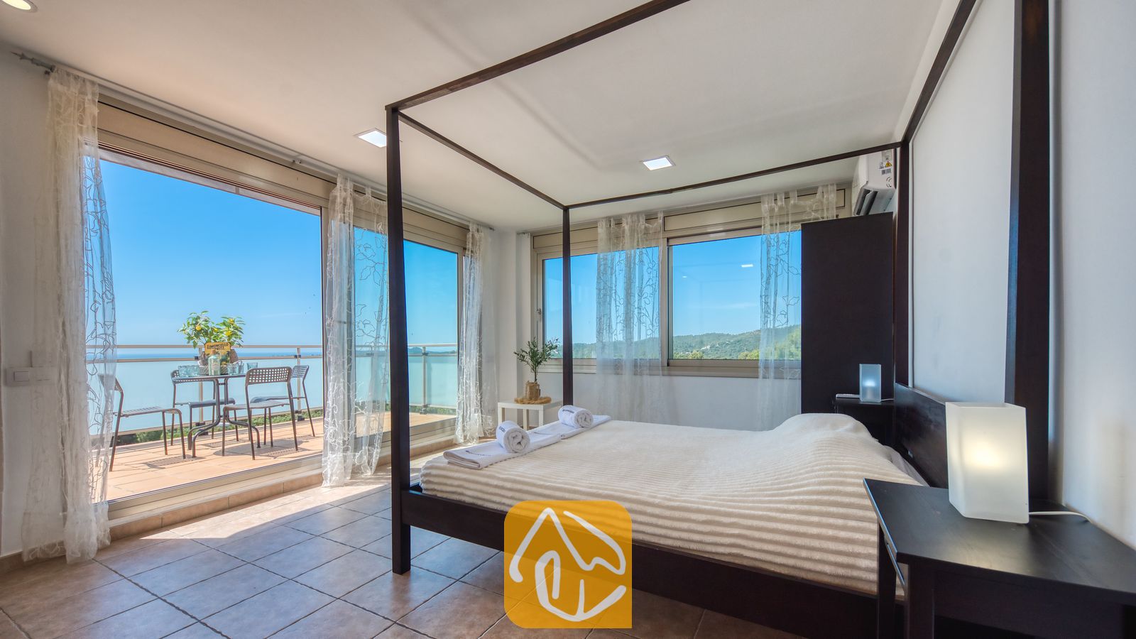Vakantievilla's Costa Brava Spanje - Villa Sandy Beach - Slaapkamer met een hemelbed van donker hout en wit linnen in Villa Sandy Beach, voorzien van glazen schuifdeuren die uitkomen op een terras met uitzicht op zee.