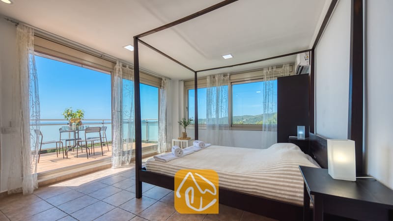 Villas de vacances Costa Brava Espagne - Villa Sandy Beach - Chambre dotée d'un lit à baldaquin en bois sombre et de linge de maison blanc à la Villa Sandy Beach, agrémentée de baies vitrées coulissantes ouvrant sur une terrasse avec vue sur la mer.