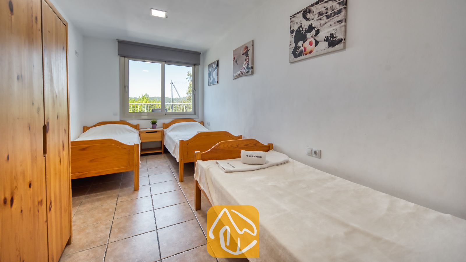 Vakantievilla's Costa Brava Spanje - Villa Sandy Beach - Slaapkamer in Villa Sandy Beach, voorzien van drie houten eenpersoonsbedden, een bijpassende kledingkast en een tegelvloer. Het raam biedt een prachtig uitzicht op het omliggende groen.