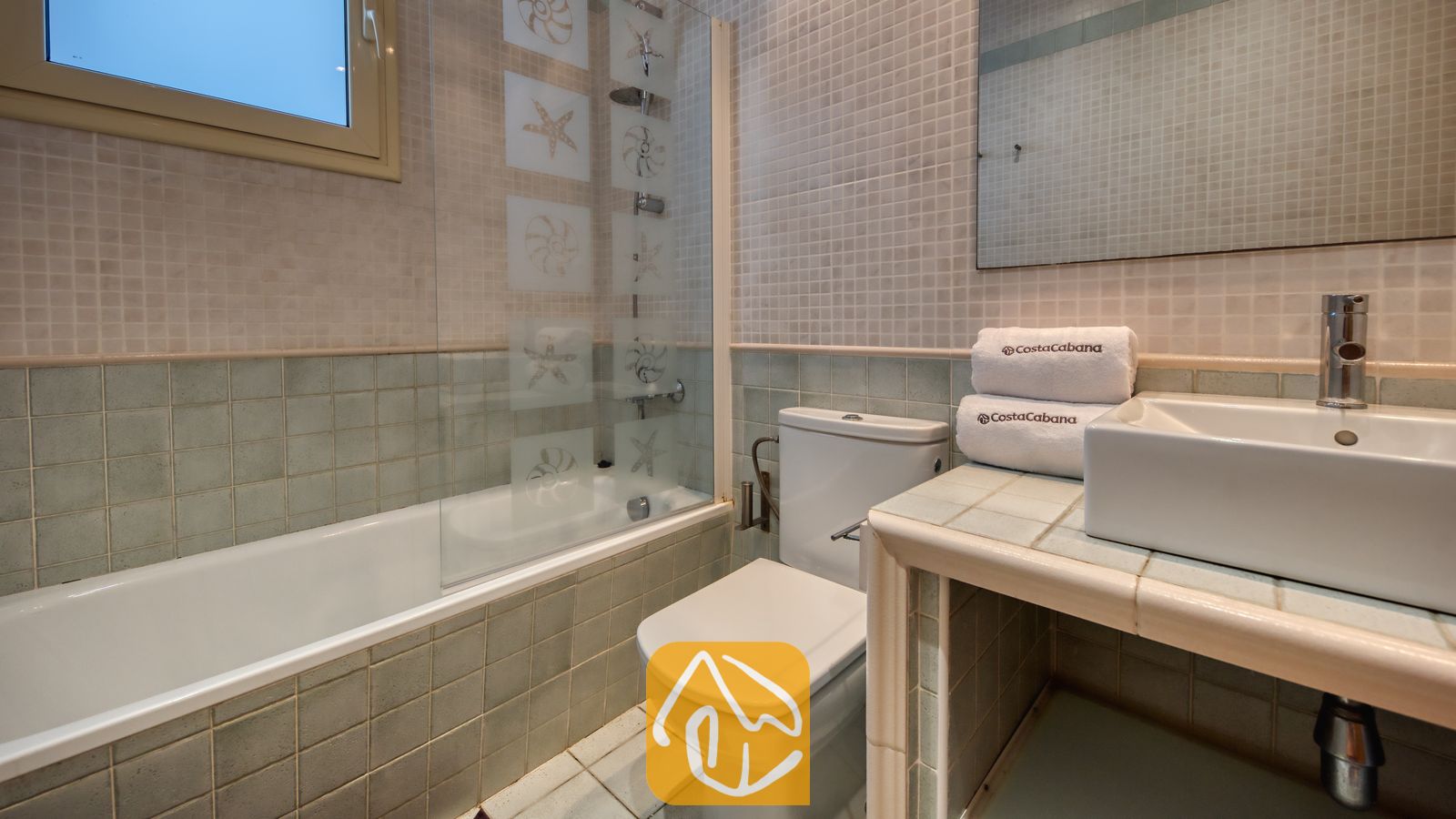 Vakantievilla's Costa Brava Spanje - Villa Sandy Beach - Badkamer met ligbad voorzien van een glazen douchescherm met zeesterpatroon, lichtgroene mozaïektegels en een witte opzetwastafel op een betegeld wastafelmeubel.