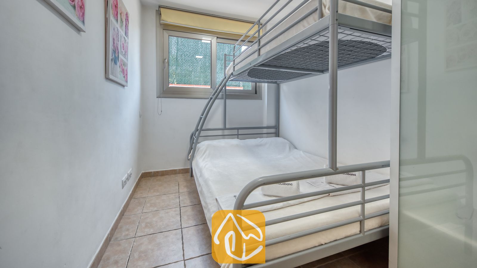 Vakantievilla's Costa Brava Spanje - Villa Sandy Beach - Slaapkamer met grijze metalen stapelbedden en wit beddengoed in Villa Sandy Beach. De ruimte is voorzien van beige plavuizen en een raam met een groen privacyscherm.