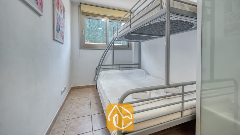 Villas de vacances Costa Brava Espagne - Villa Sandy Beach - Chambre équipée de lits superposés en métal gris et de linge de maison blanc à la Villa Sandy Beach. L'espace est doté d'un sol en carrelage beige et d'une fenêtre agrémentée d'un brise-vue vert.