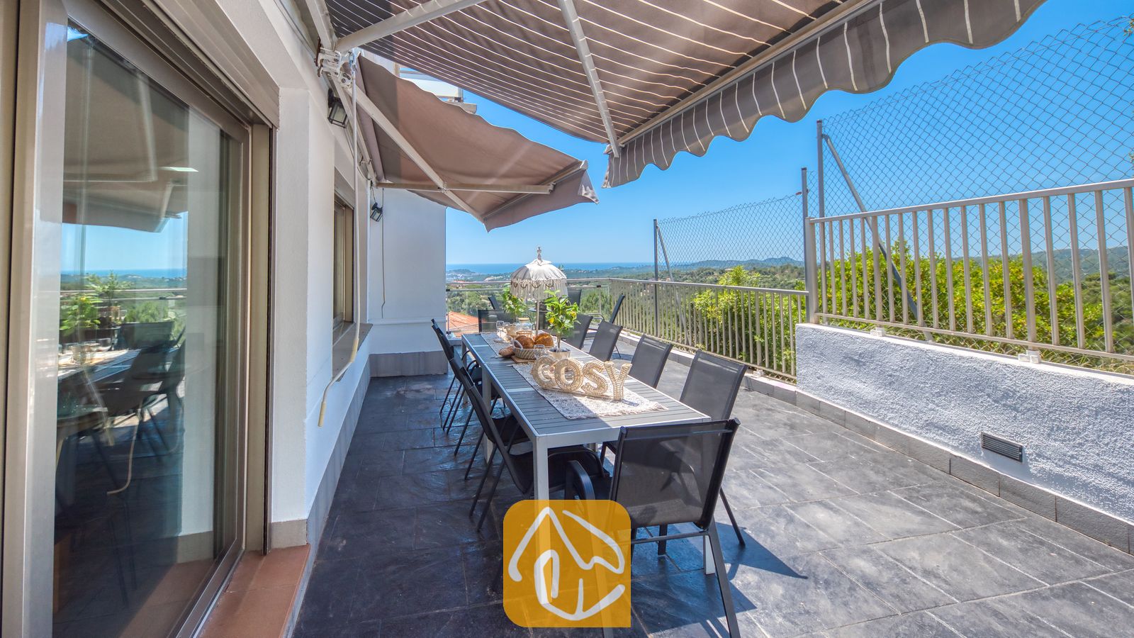 Vakantievilla's Costa Brava Spanje - Villa Sandy Beach - Terras bij Villa Sandy Beach met een lange eettafel, zwarte mesh-stoelen en uitschuifbare gestreepte zonneschermen met uitzicht op de zee en de heuvels.