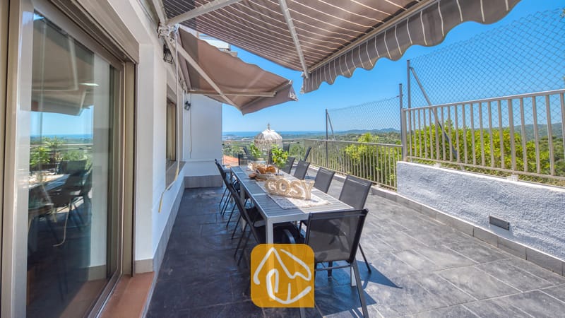 Villas de vacances Costa Brava Espagne - Villa Sandy Beach - Terrasse de la Villa Sandy Beach dotée d'une longue table de salle à manger, de chaises en maille noire et de stores rayés rétractables, offrant une vue imprenable sur la mer et les collines.